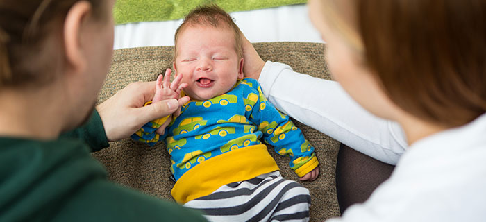 A baby is on a blanket and two adults are standing on the side.