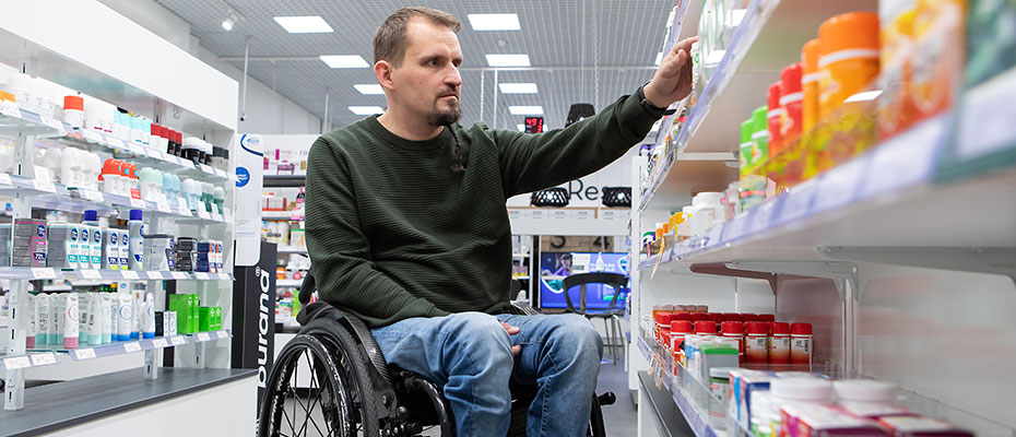 Pyörätuolissa istuva asiakas etsii lääkettä apteekin hyllyltä.