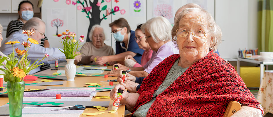 Elderly people making arts and crafts around a table. An elderly lady in the front is looking straight at the camera.