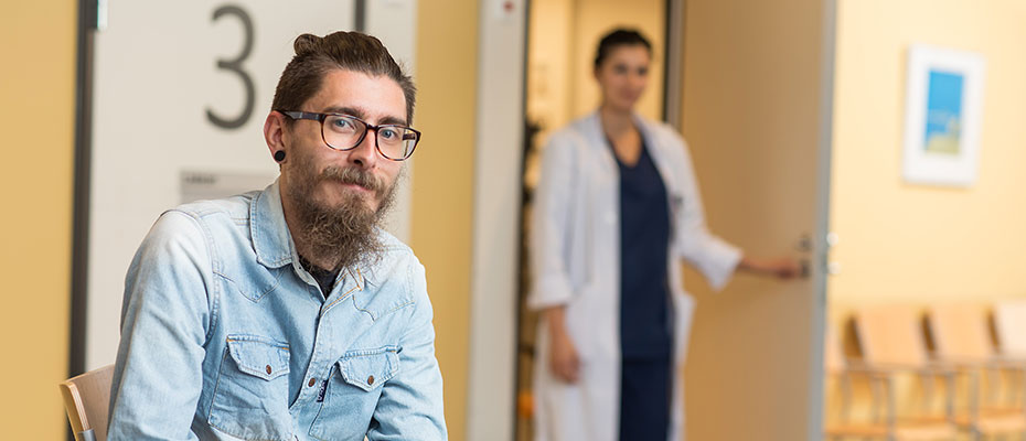 A man is sitting in a waiting room facing the camera. In the background is a wellbeing services county employee.