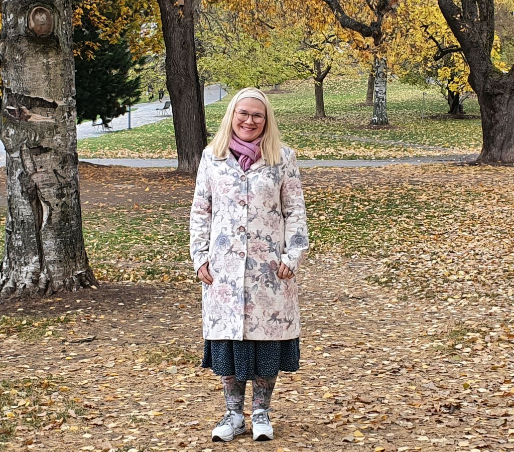 Nainen seisoo puistossa nurmikolla puiden välissä ja katsoo nauraen kameraan. Nurmikolla on paljon puista pudonneita lehtiä. Taustalla näkyy puiston hiekkateitä.