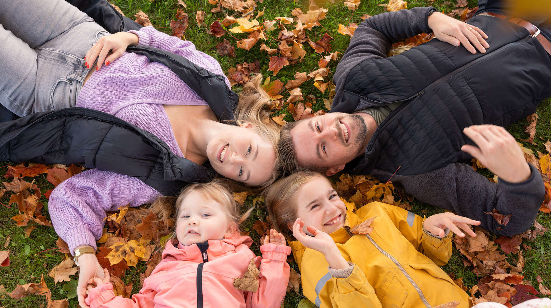 Kaksi aikuista ja kaksi tyttöä makaa selällään ruskalehtien päällä maassa niin, että heidän päänsä koskettavat toisiaan.