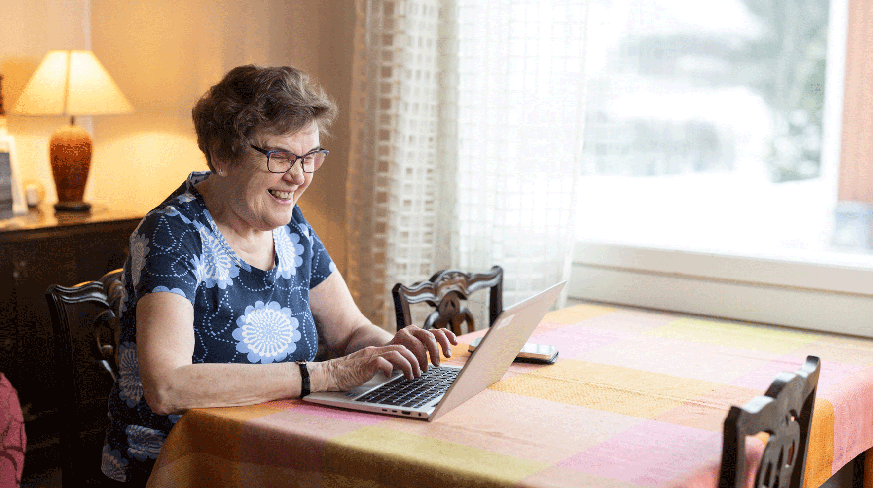Iloinen iäkäs nainen istuu keittiön pöydän ääressä tietokoneella. Hänellä sininen kukikas paita ja ruskea lyhyt tukka. 
