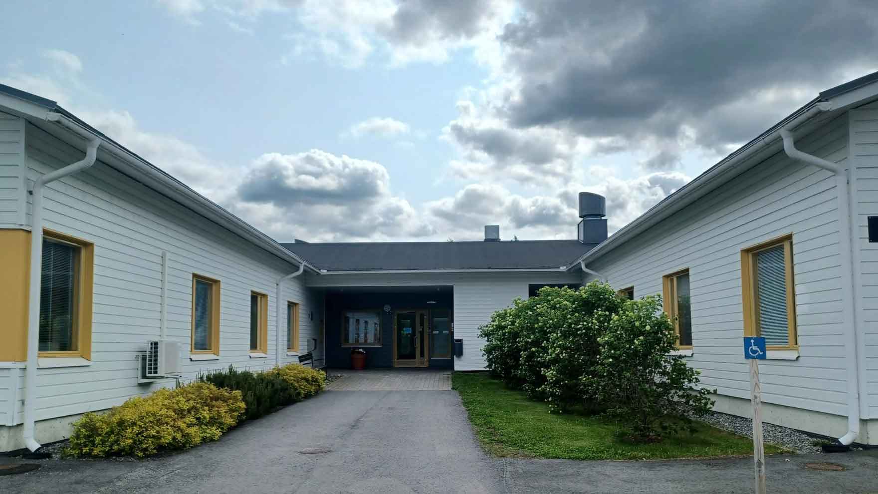 Vaaleasta laudasta tehty matala rakennus ja sen sisäänkäynti, jossa istutuksia