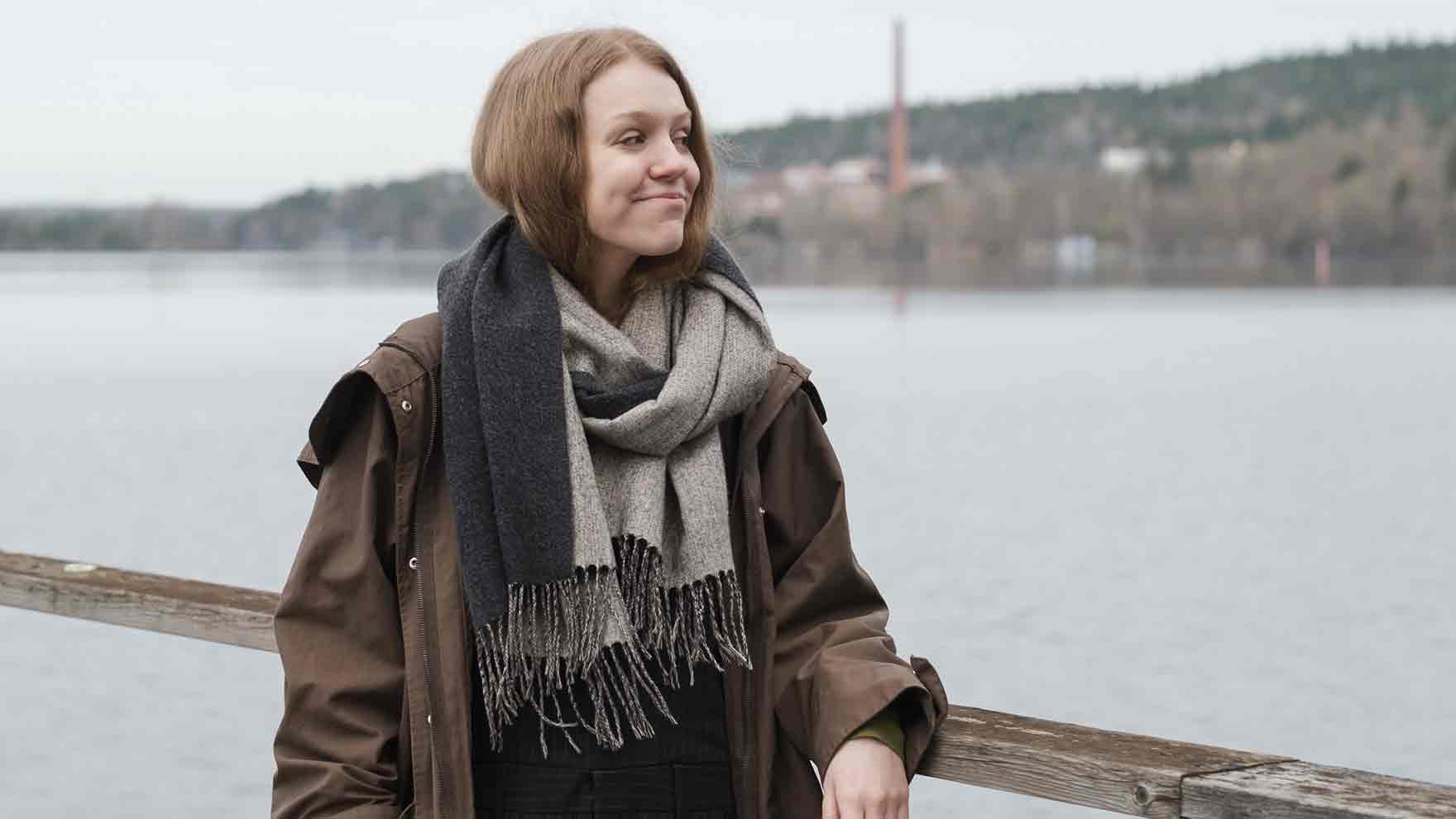 Pitkähiuksinen Alisa nojaa kaiteeseen järven rannalla. Hän katsoo sivulle ja hymyilee. 