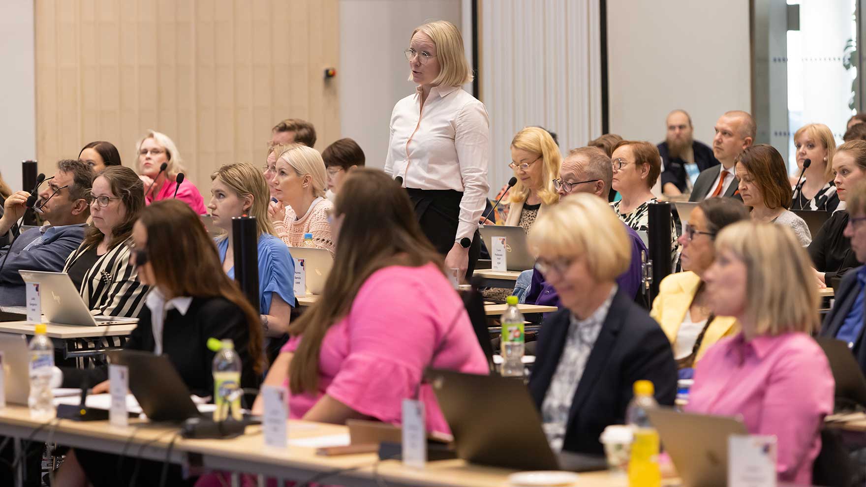 Aluevaltuutetut valtuuston kokouksessa. Yksi valtuutetuista seisoo ja käyttää puheenvuoroa.