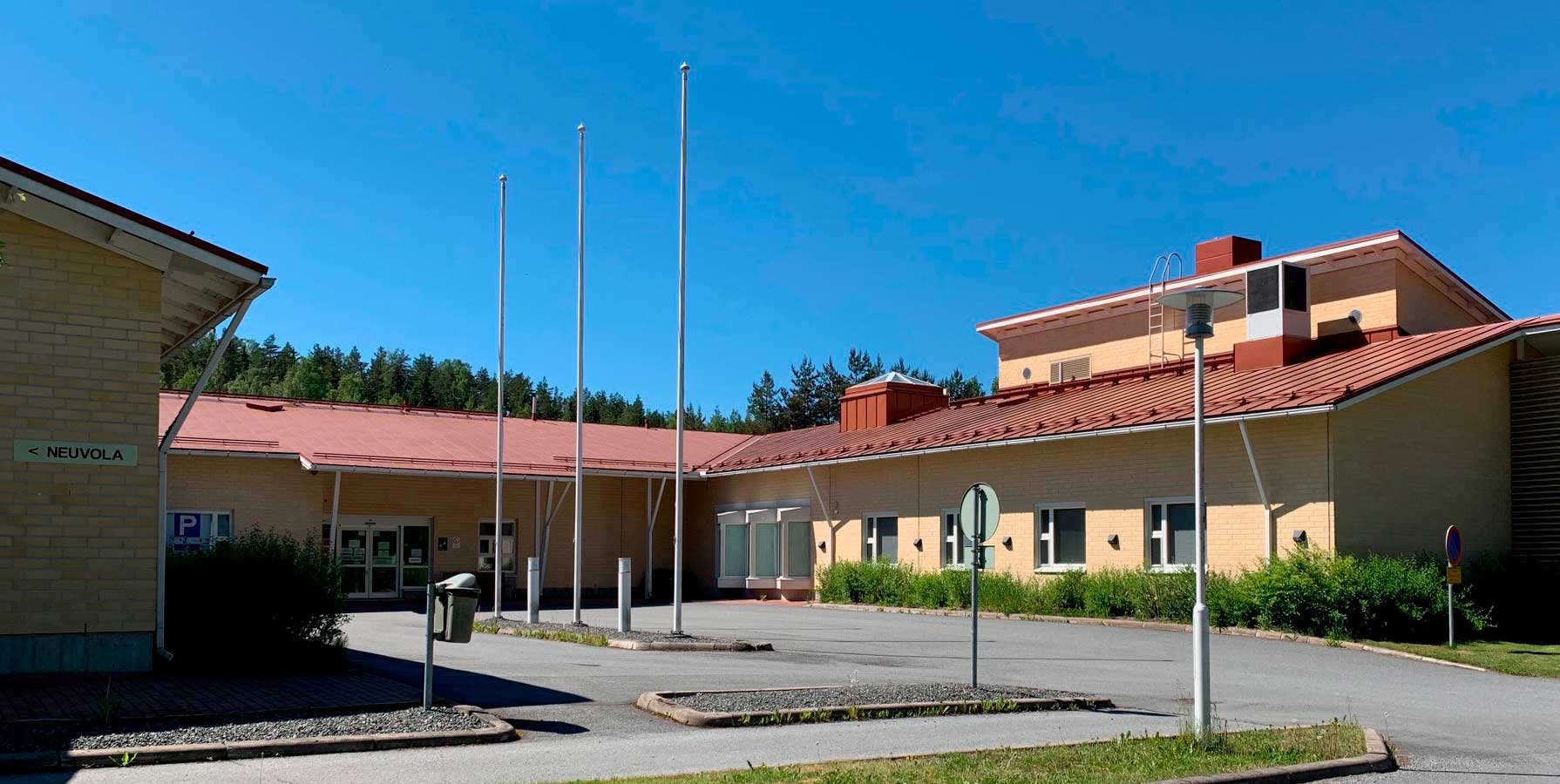 Yksikerroksinen, U-kirjaimen muotoinen, keltatiilinen rakennus, jonka pihalla on kolme lipputankoa. 