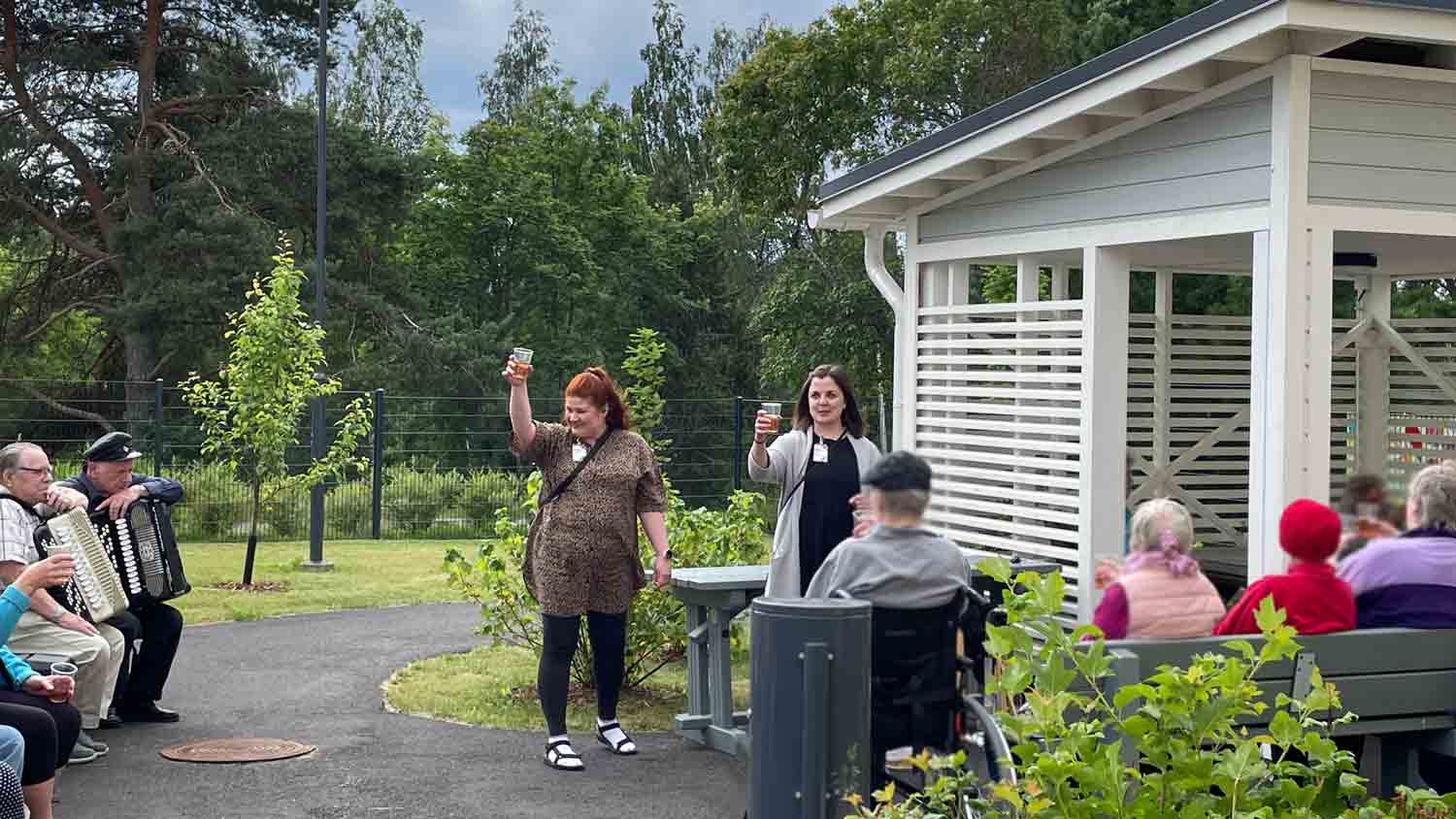 Kultturiohjaajat skoolaavat juomalla Höllinkodon pihalla. Ympärillä istuu asukkaita ja tilaisuuden vieraita. 