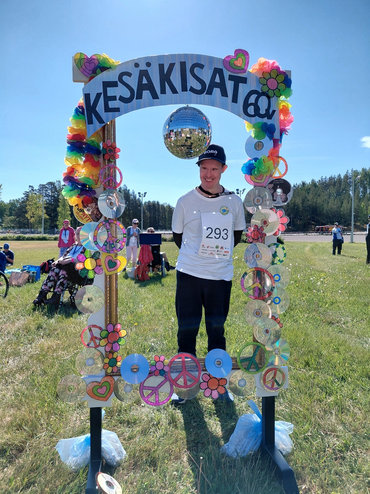 Henkilö poseeraa Kesäkisat 60 v. -kaaren takaa. Henkilöllä on paitaan kiinnitettynä kilpailunumero 293..
