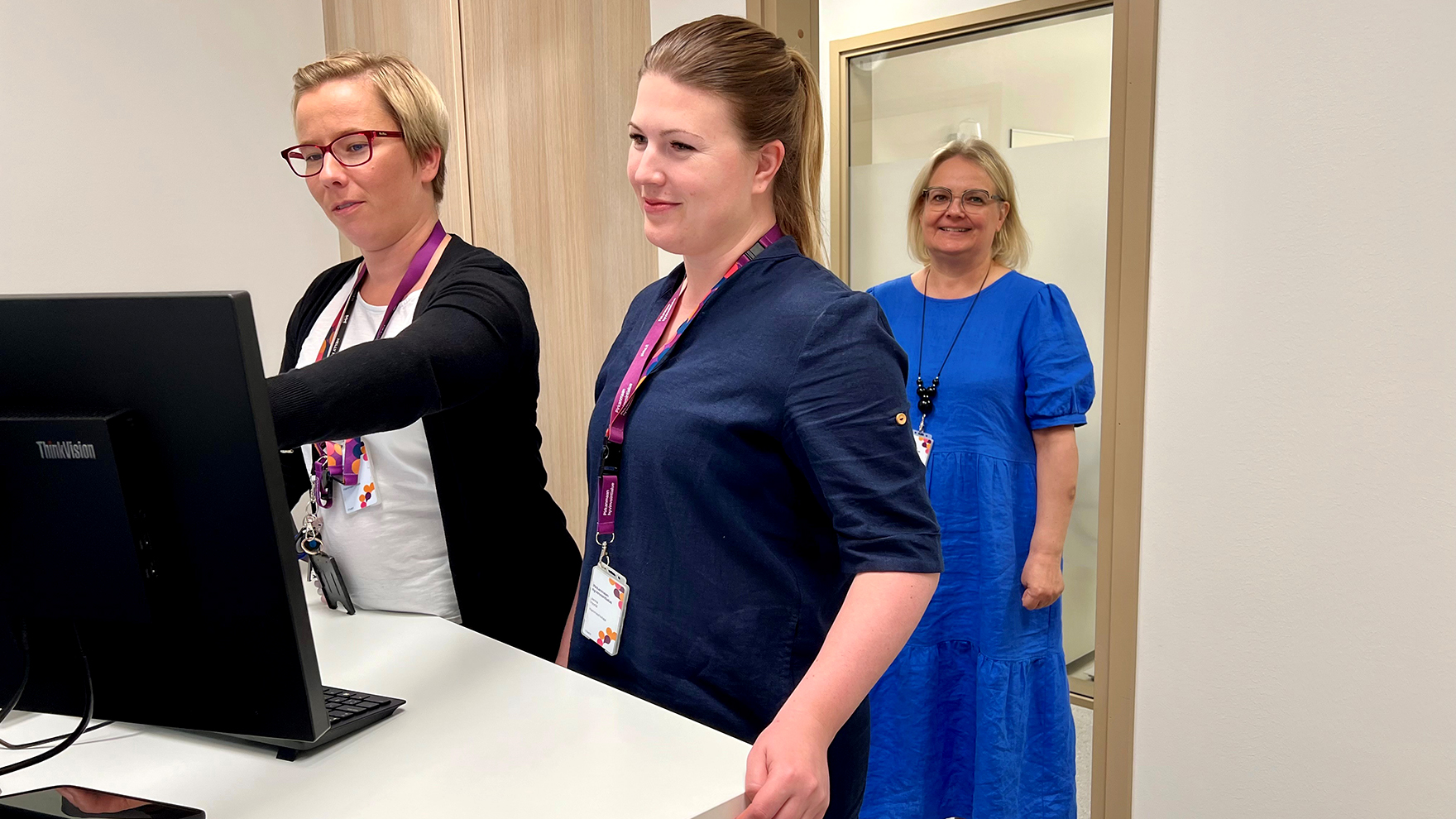 Kaksi naista seisoo työpöydän ääressä ja katsoo tietokeen näyttöä. Takana ovella seisoo yksi nainen sinisessä mekossa ja hymyilee.