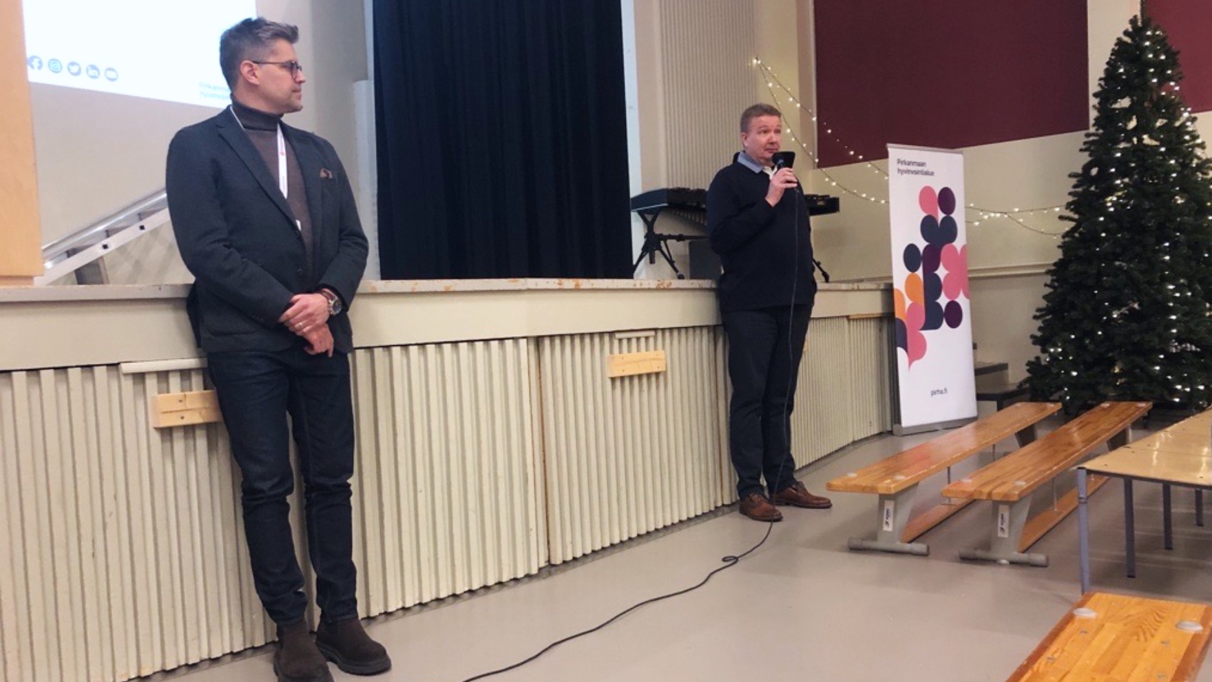 Kaksi miestä seisoo juhlasalissa lavan edessä puhumassa yleisölle.