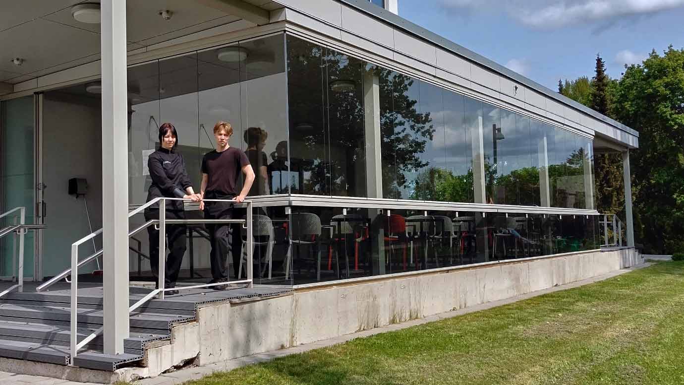 Nuori nainen ja mies seisovat lasiterassilla. Terassilla näkyy tyhjiä tuoleja ja pöytiä.