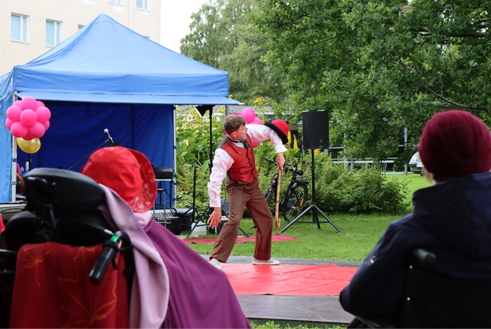 Mieshenkilö pitää kädessään kävelykeppiä ja kyynärpäässään punaista hattua. Mies on puisen alustan päällä, taustalla näkyy sininen katos, kaiutin ja vehreää luontoa.