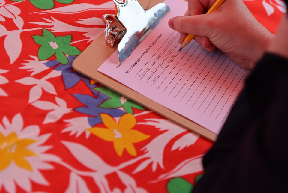 Henkilö kirjoittaa kirjoitusalustaa vasten paperille muistoistaan. Kirjoitusalusta on kukikkaalla, punaisella pöytäliinalla.