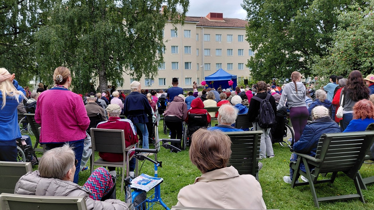 Yleisö katselee kohti sinistä katosta, jossa esiintyjä esiintyy. Ympärillä on vihreitä puita ja taustalla näkyy vaaleankeltainen rakennus.