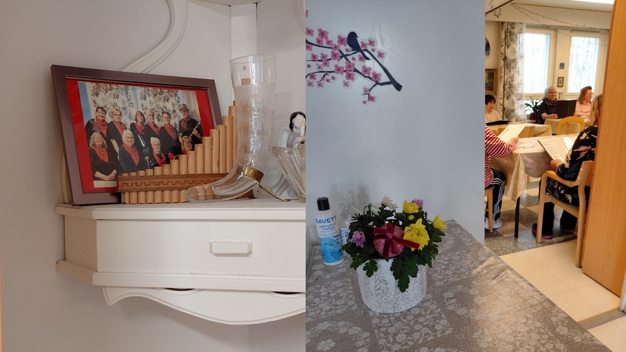 Vasemmalla on hyllyn päällä valokuva kehystettynä ja koriste-esineitä. Oikealla kuvassa edessä on pöydällä kukkapurkki ja taaempana näkyy kuoron harjoitukset.