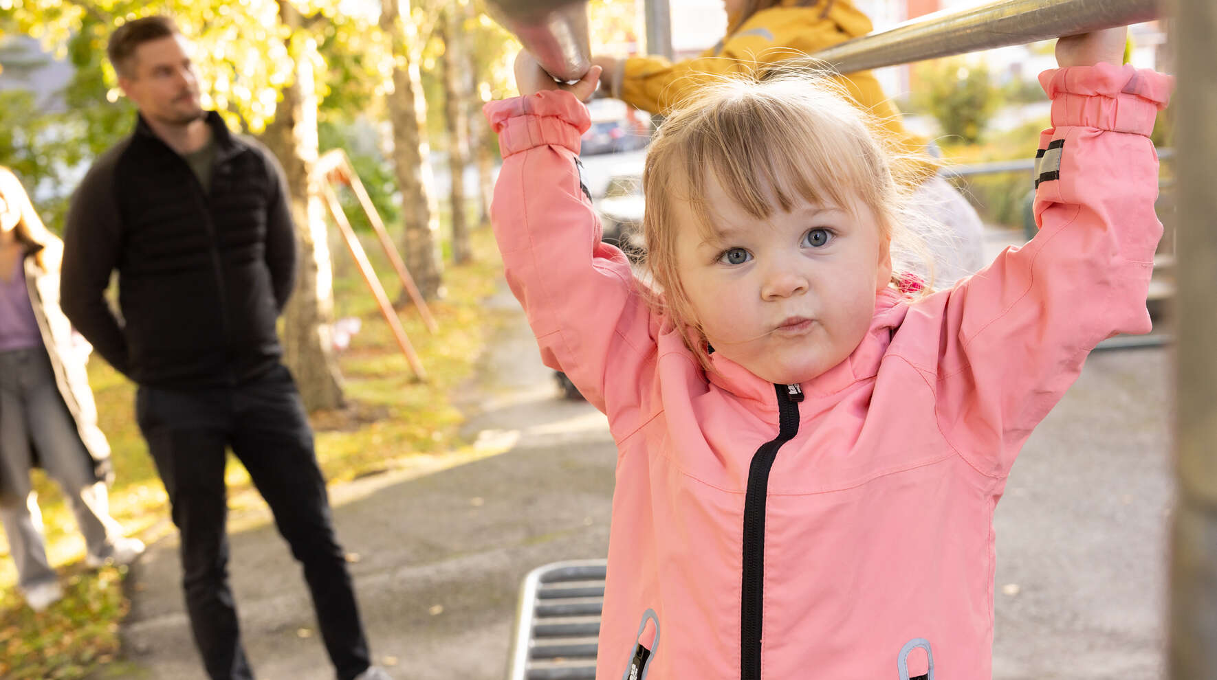 Kaksi aikuista seuraa kahden lapsen kiipeilyä mattotelineellä. Etualalla oleva pieni tyttö katsoo kohti suu supussa.