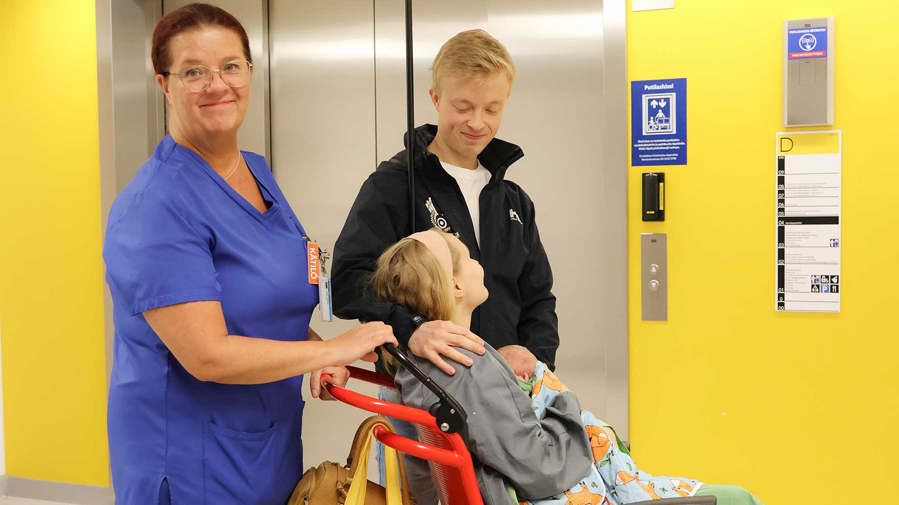 Kätilö työntää vauvaa sylissään pitävää synnyttänyttä äitiä pyörätuolilla hissiin. Mukana on mysö vauvan isä.