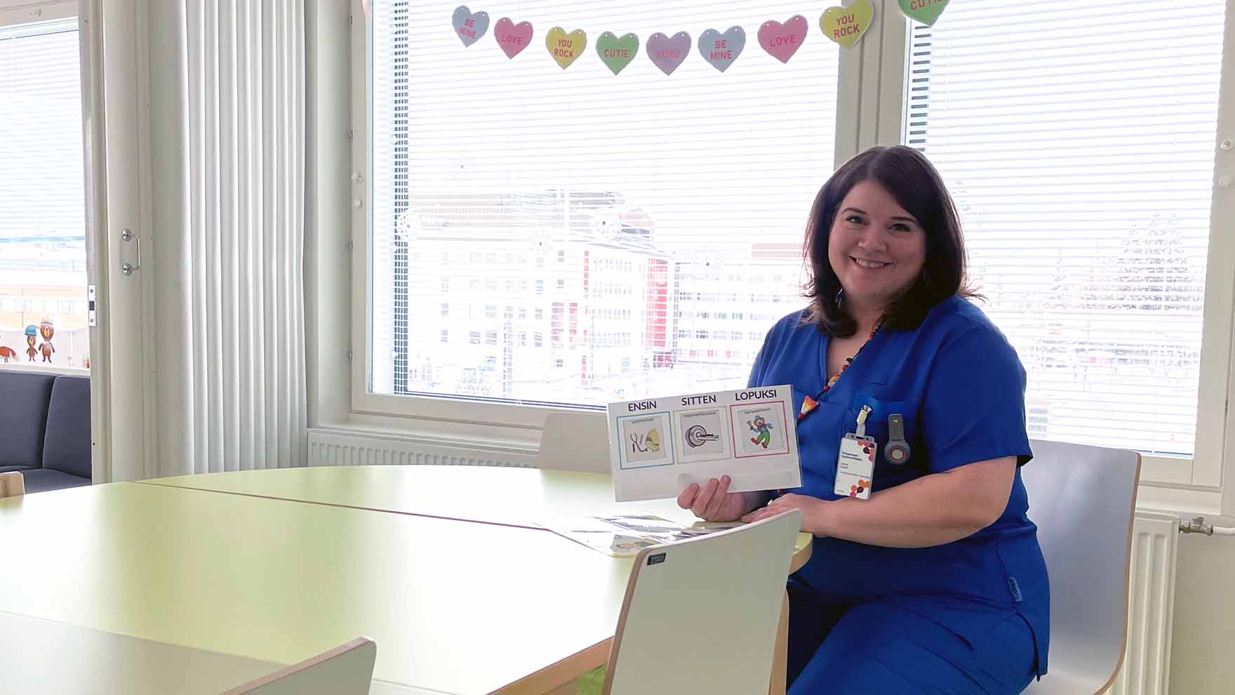 Hymyilevä nainen istuu pöydän ääressä ja esittelee vuorovaikutuksen tukena käytettävää kuvakorttia.