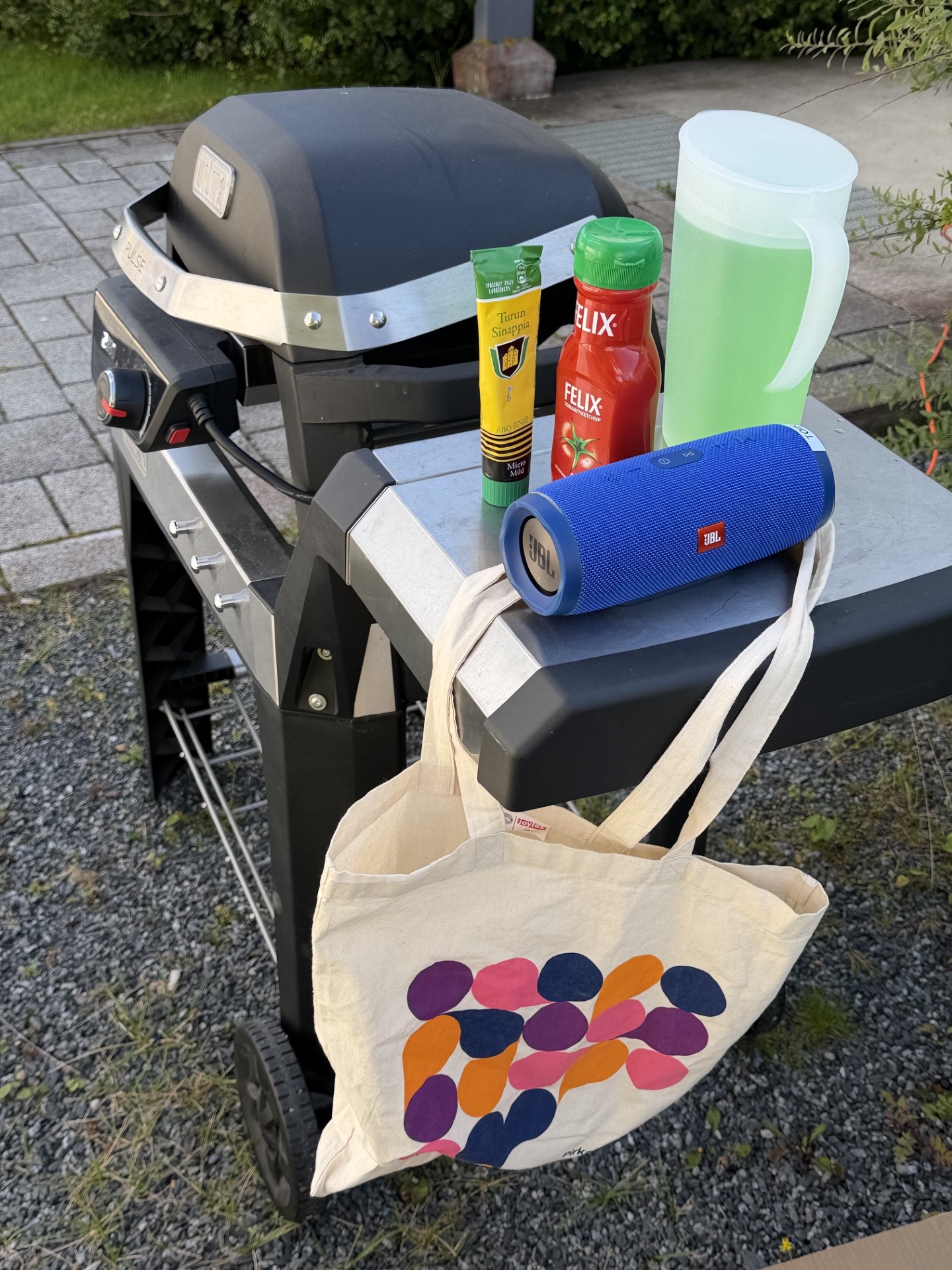Grilli, jonka sivupöydällä on kaiutin, ketsuppia, sinappia ja mehukannu. Sivutason kulmasta roikkuu Pirhan kangaskassi.