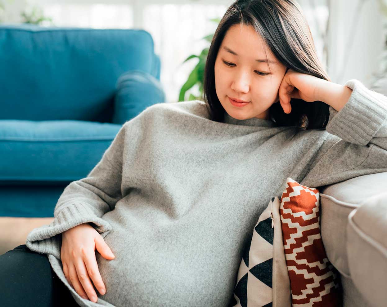 Odottava nainen istuu sohvalla ja pitää toista kättään vatsansa päällä.