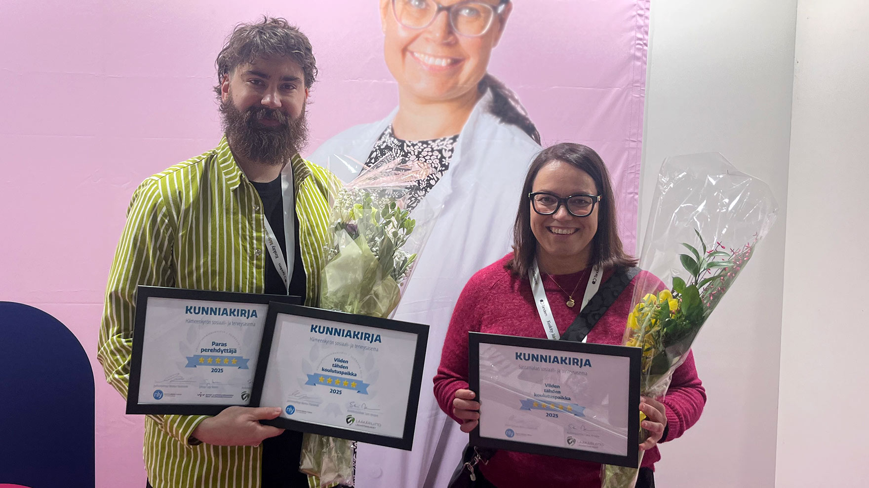 Kaksi henkilöä seisovat hymyillen vierekkäin. Toisella on kädessään kaksi kunniakirjaa, toisella yksi.