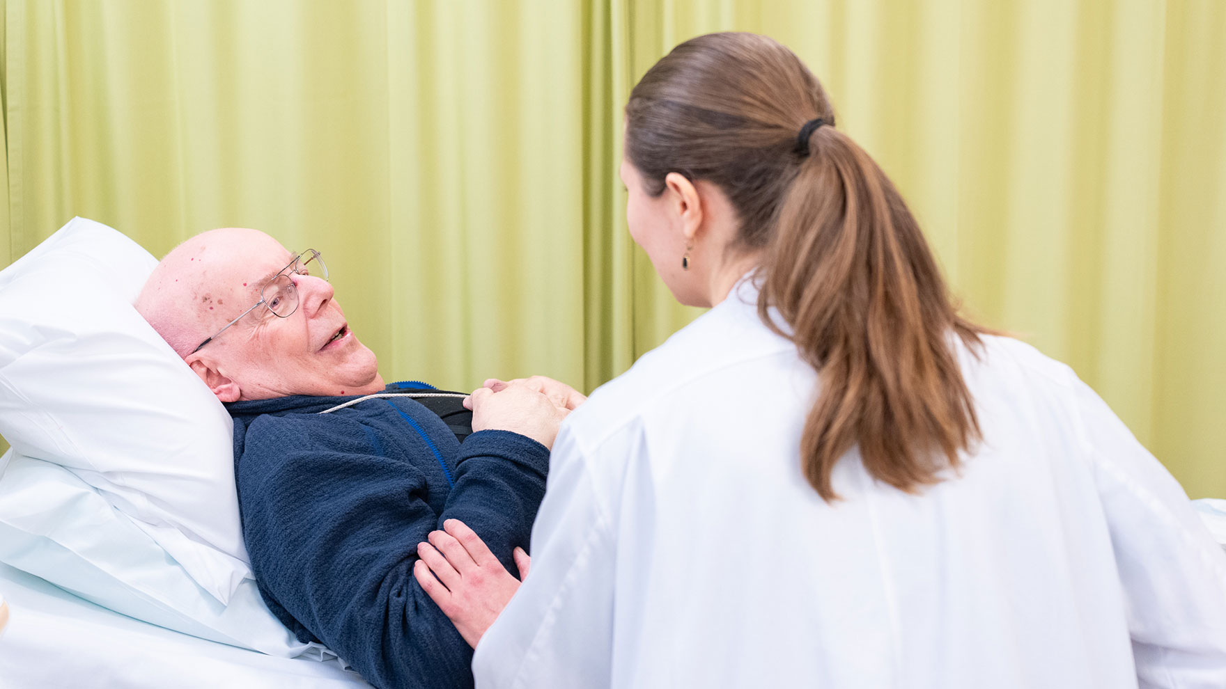 Terveydenhuollon ammattilainen juttelee sängyssä makaavan potilaan kanssa.
