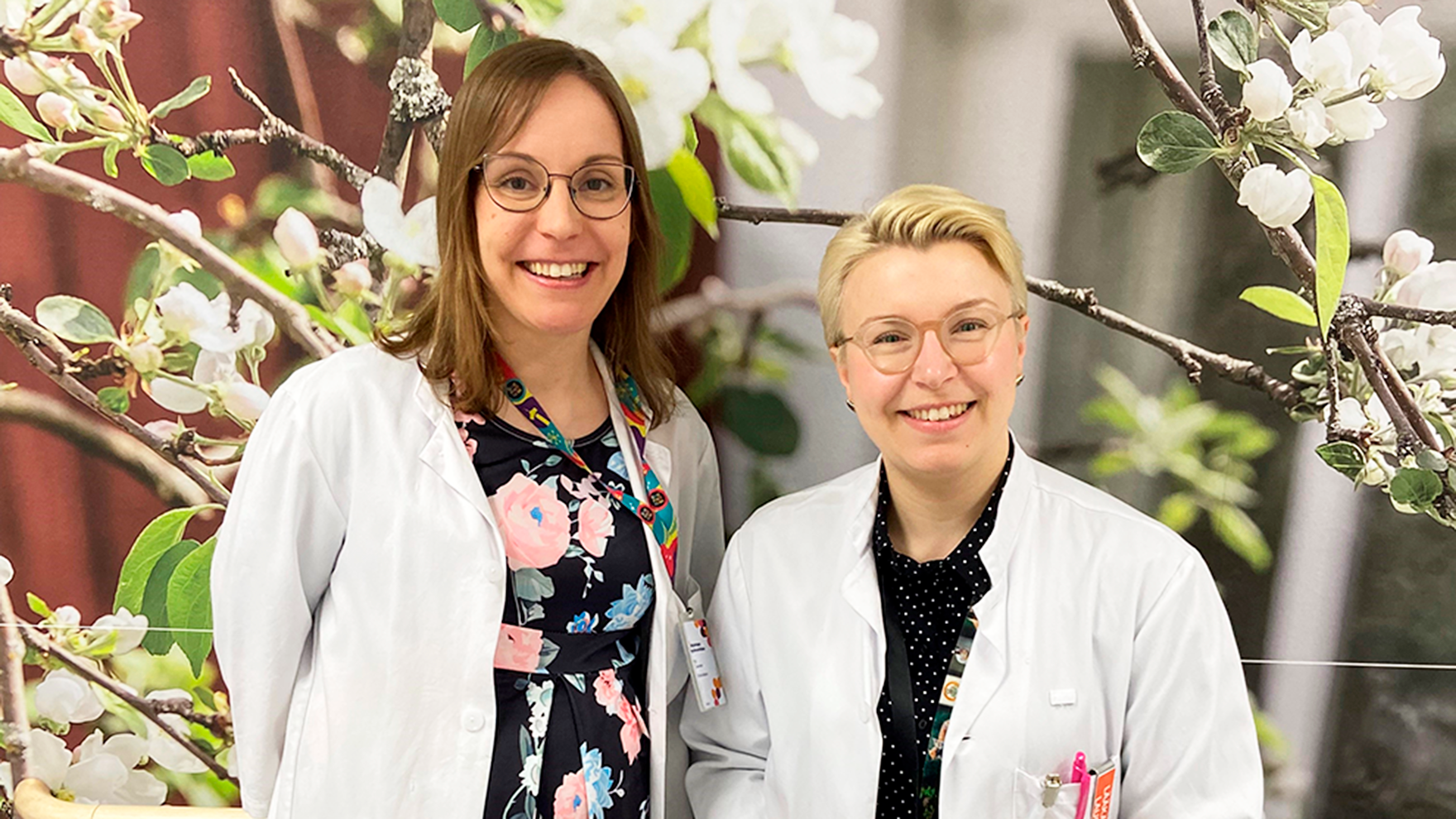 Kuvassa ovat lääkärintakki yllään Elli Toivonen ja Laura Seppälä. Taustalla näkyy valokuvatapetti, jossa on valkoisenaan kukkivia oksia.