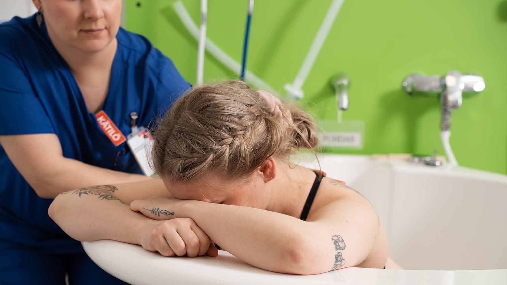 Synnyttäjä istuu ammeessa ja nojaa päätään ammeen reunaan. Kätilö on ammeen takana tukena.