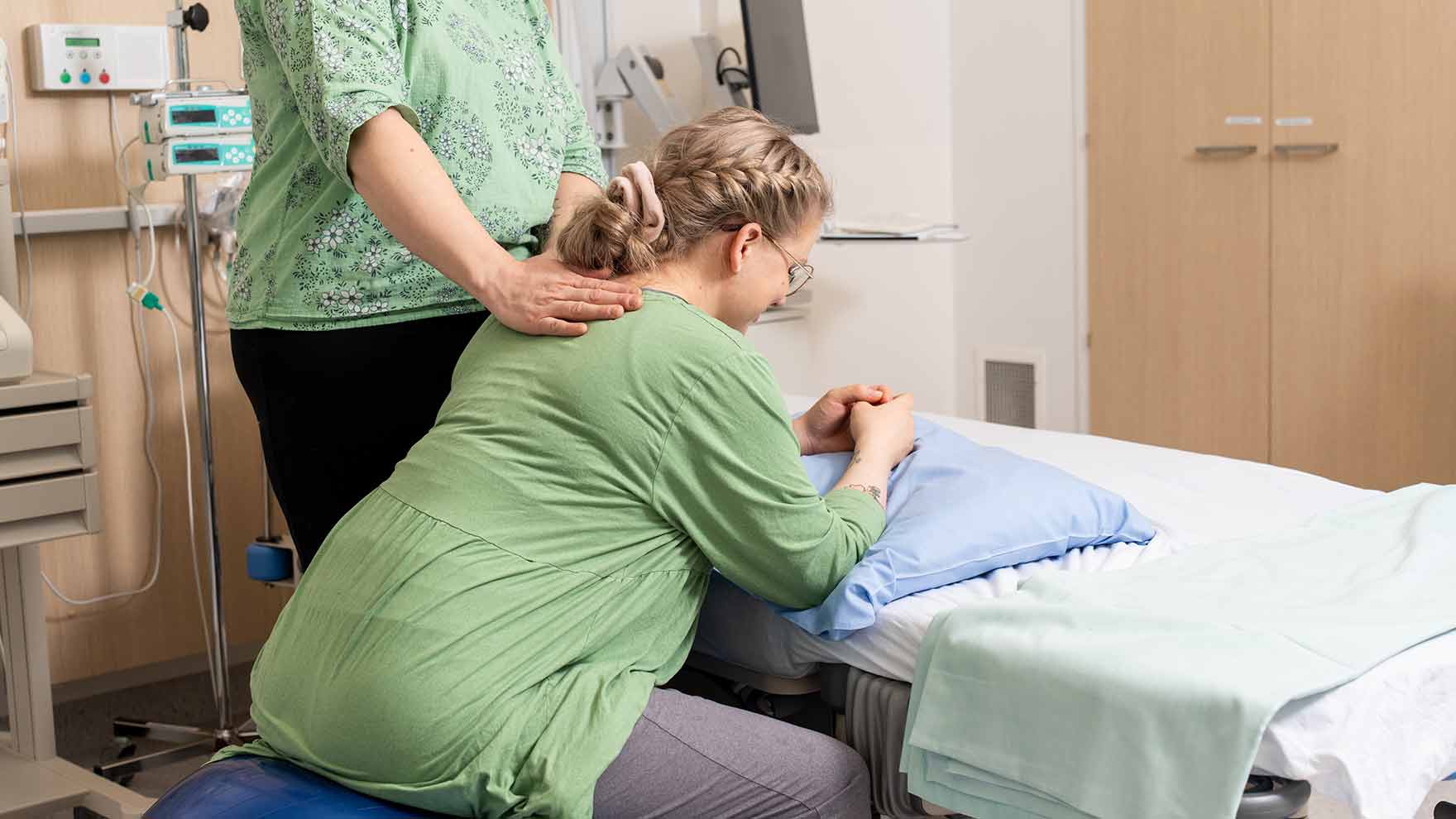 Synnyttäjä nojaa kumipallon päällä sänkyä kohti. Hänen vierellään seisoo tukihenkilö käsi synnyttäjän olkapäällä.