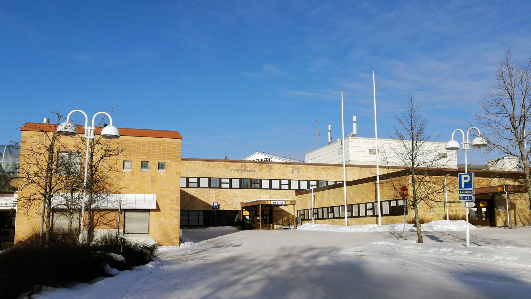 Valkeakosken sosiaali- ja terveysaseman keltatiilinen rakennus aurinkoisena talvipäivänä sinistä taivasta vasten. Etualalla on lunta.
