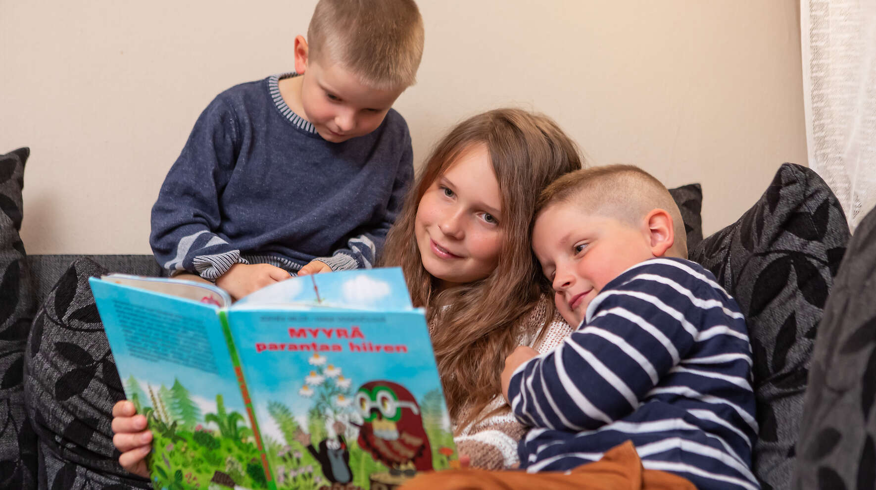 Kolme lasta istuu sohvalla lukemassa kirjaa yhdessä.