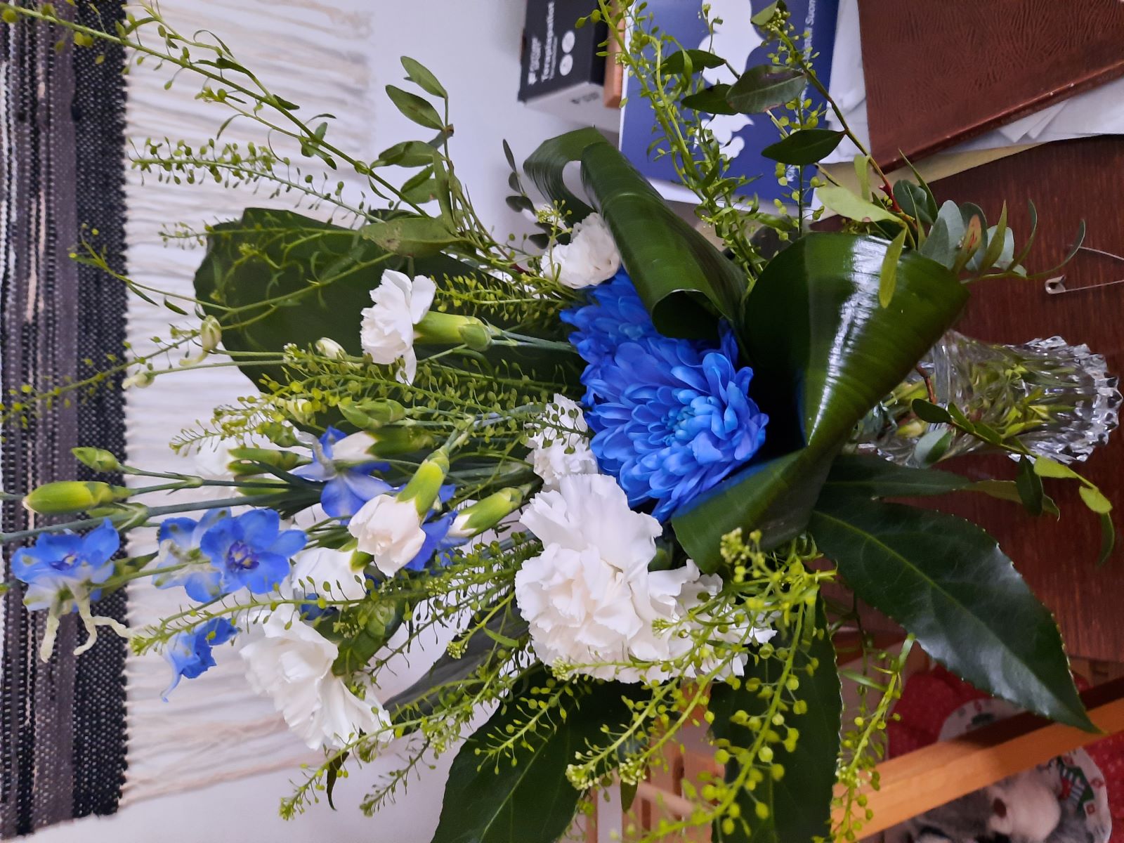 Pöydällä maljakossa on kaunis kimppu valkoisia ja sinisiä kukkia sekä vihreitä kasveja.