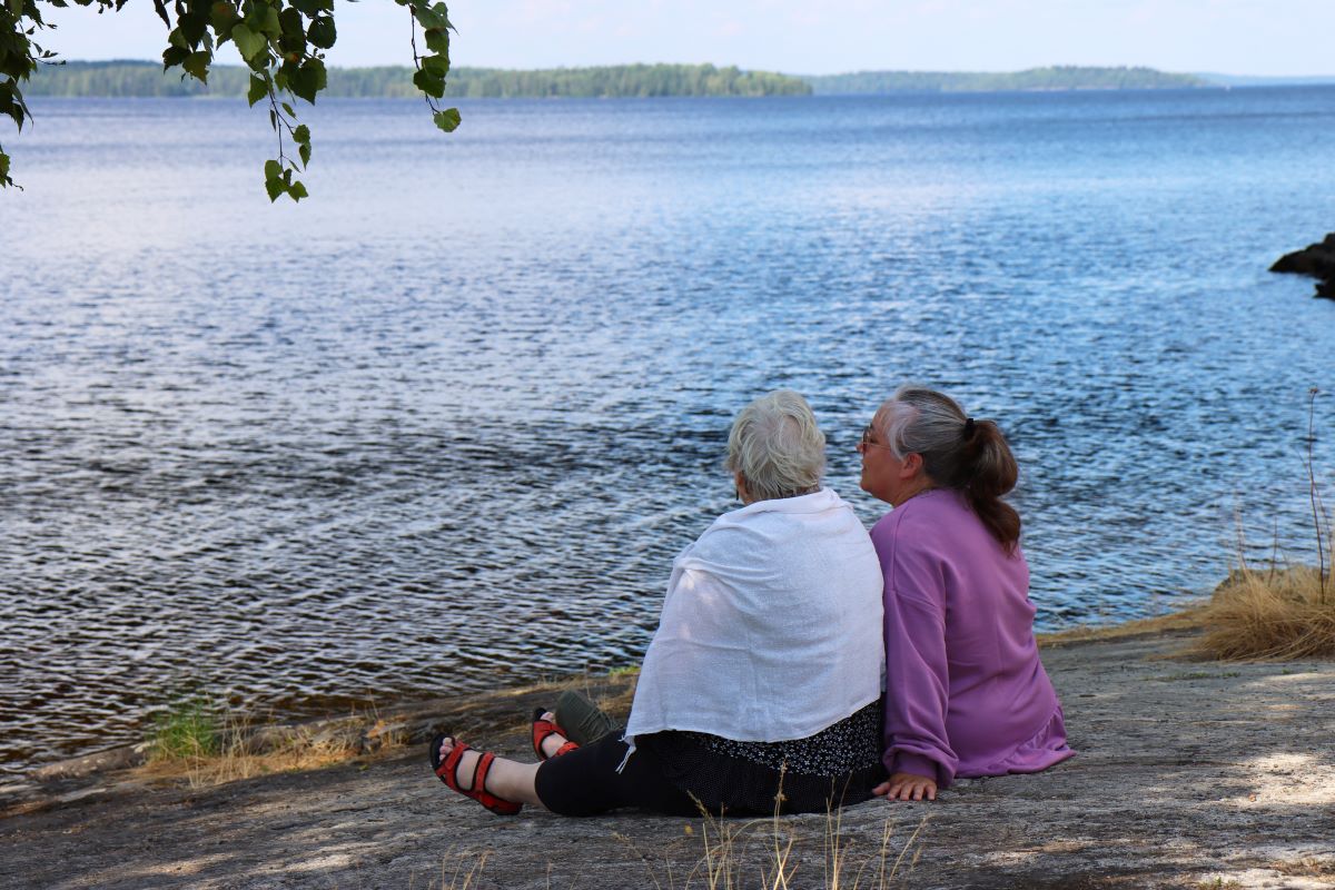 Kaksi ikääntynyttä istuvat kalliolla järven rannalla.