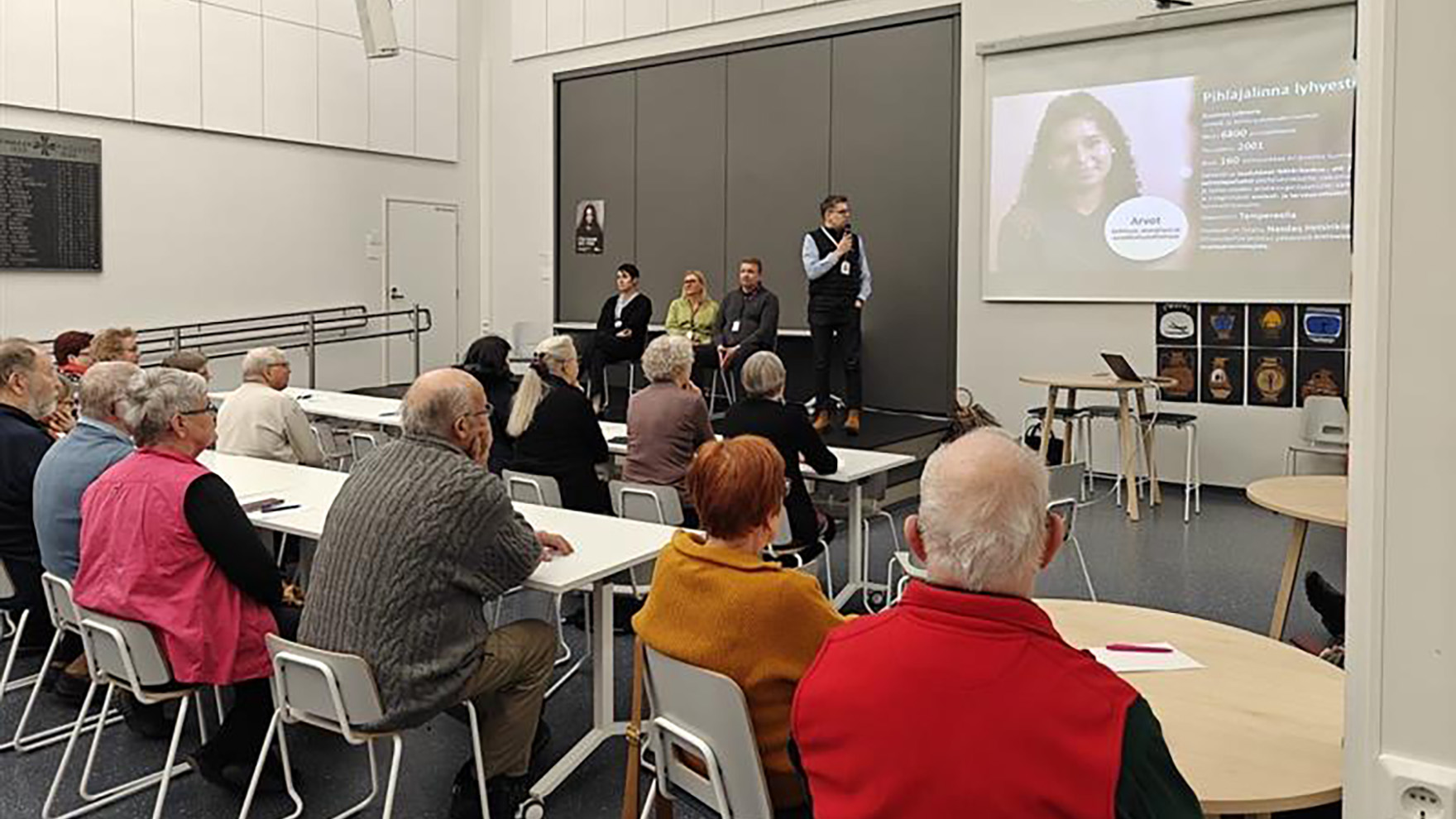 Yleisöä istumassa salissa ja lavalla puhujia istumassa. Yksi puhuja seisoo ja kertoo asioista.