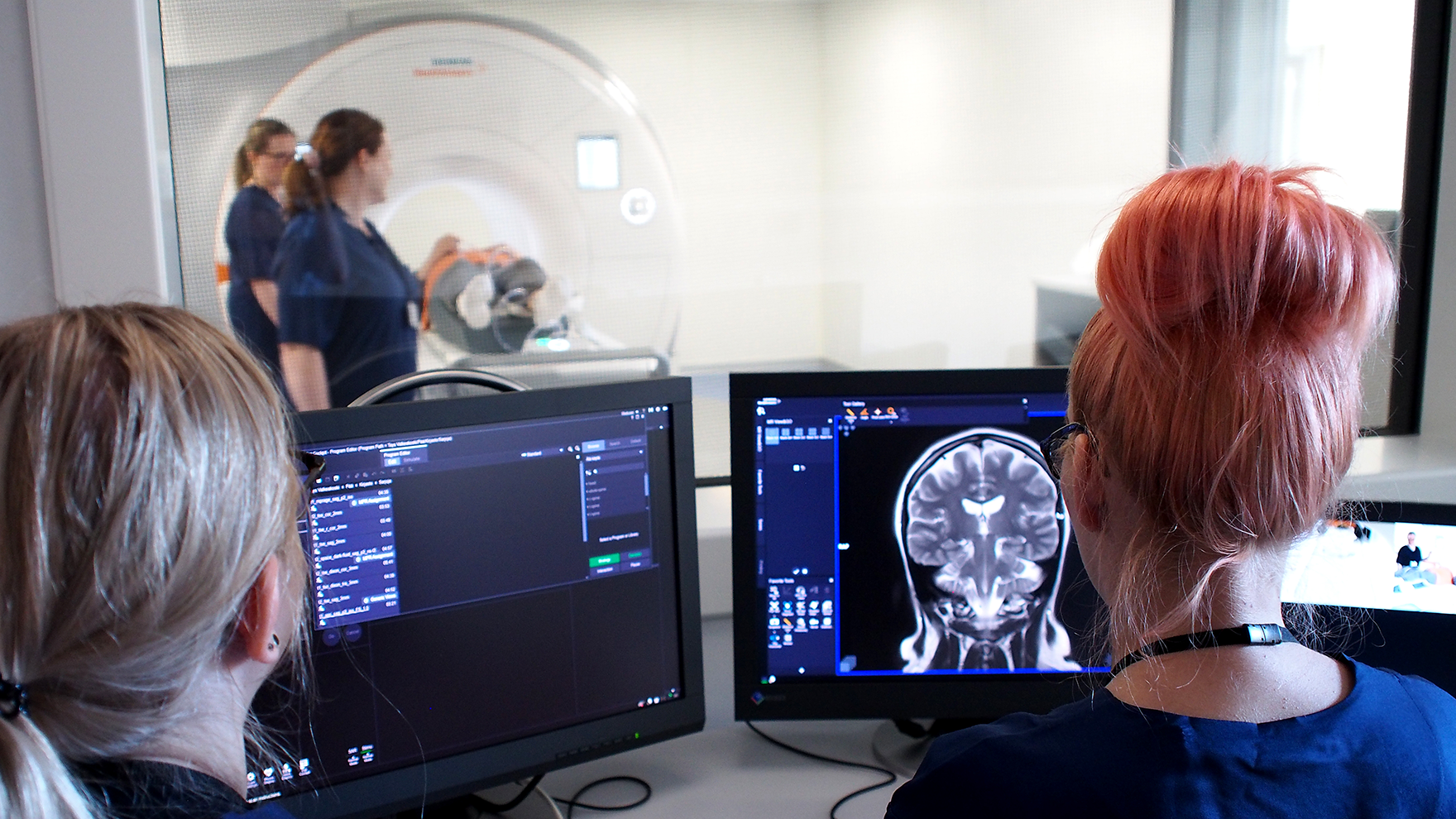 Kaksi työntekijää katsoo tietokoneelta magneettikuvia, taustalla potilas menossa tutkittavaksi kuvantamislaitteeseen.
